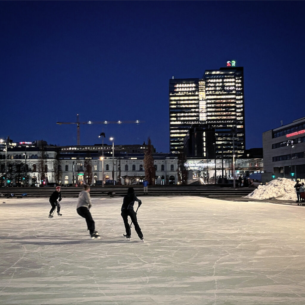 Ice skaters playing hockey on a rink downtown, with tall building in back
